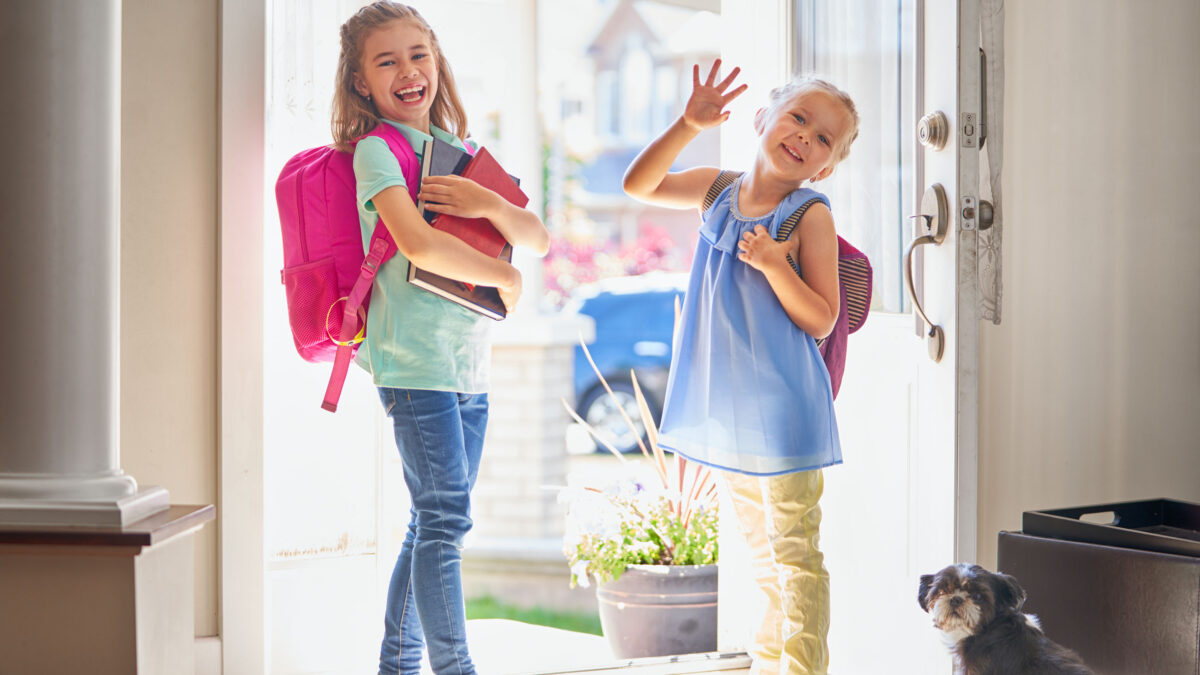 two girls with backpacks smiling and heading out the door to go to school