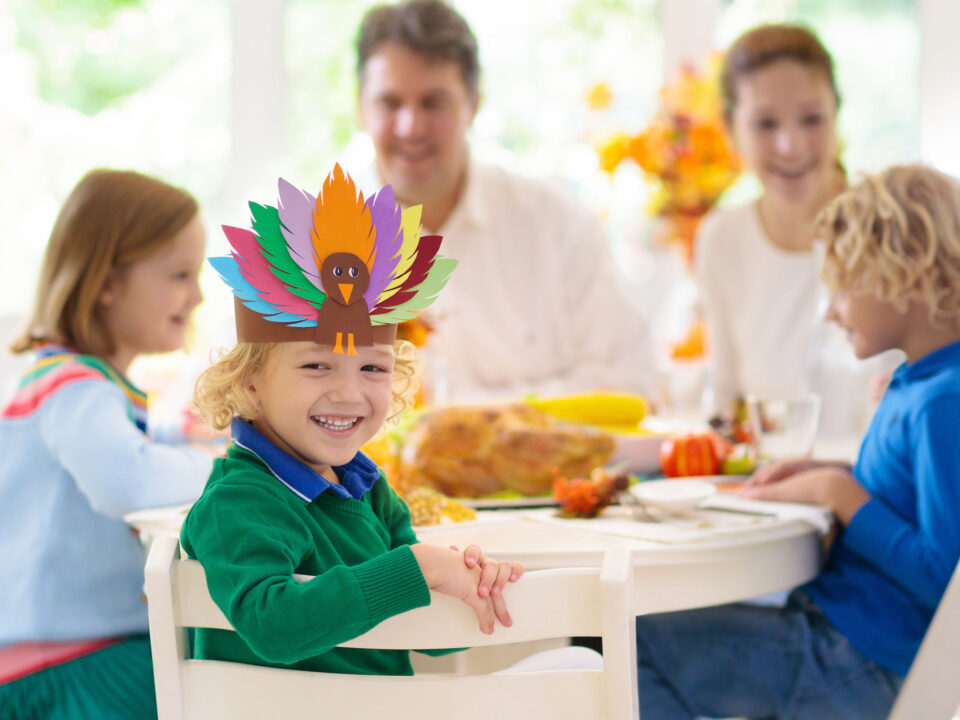 a little boy wearing a paper turkey headband and sitting around a table with his family on Thanksgiving